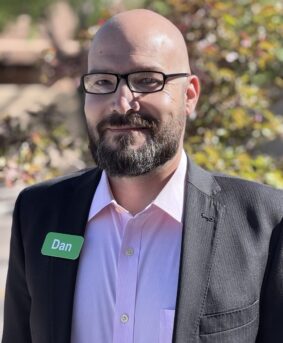 IMG_6549 Headshot of Dan Lockyer wearing a light pink shirt, black suit, and glasses outdoors.